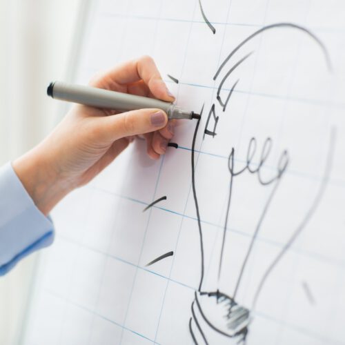 close up of hand drawing light bulb on flip chart business, people, idea, startup and education concept - close up of hand with marker drawing light bulb on flip chart at office