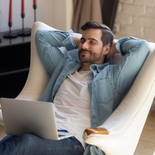 Peaceful young man daydreaming in comfortable armchair, holding computer. Peaceful young man daydreaming in comfortable armchair with computer on knees. Calm guy taking break after working with laptop, controlling fatigue or stress, resting alone in living room at home.